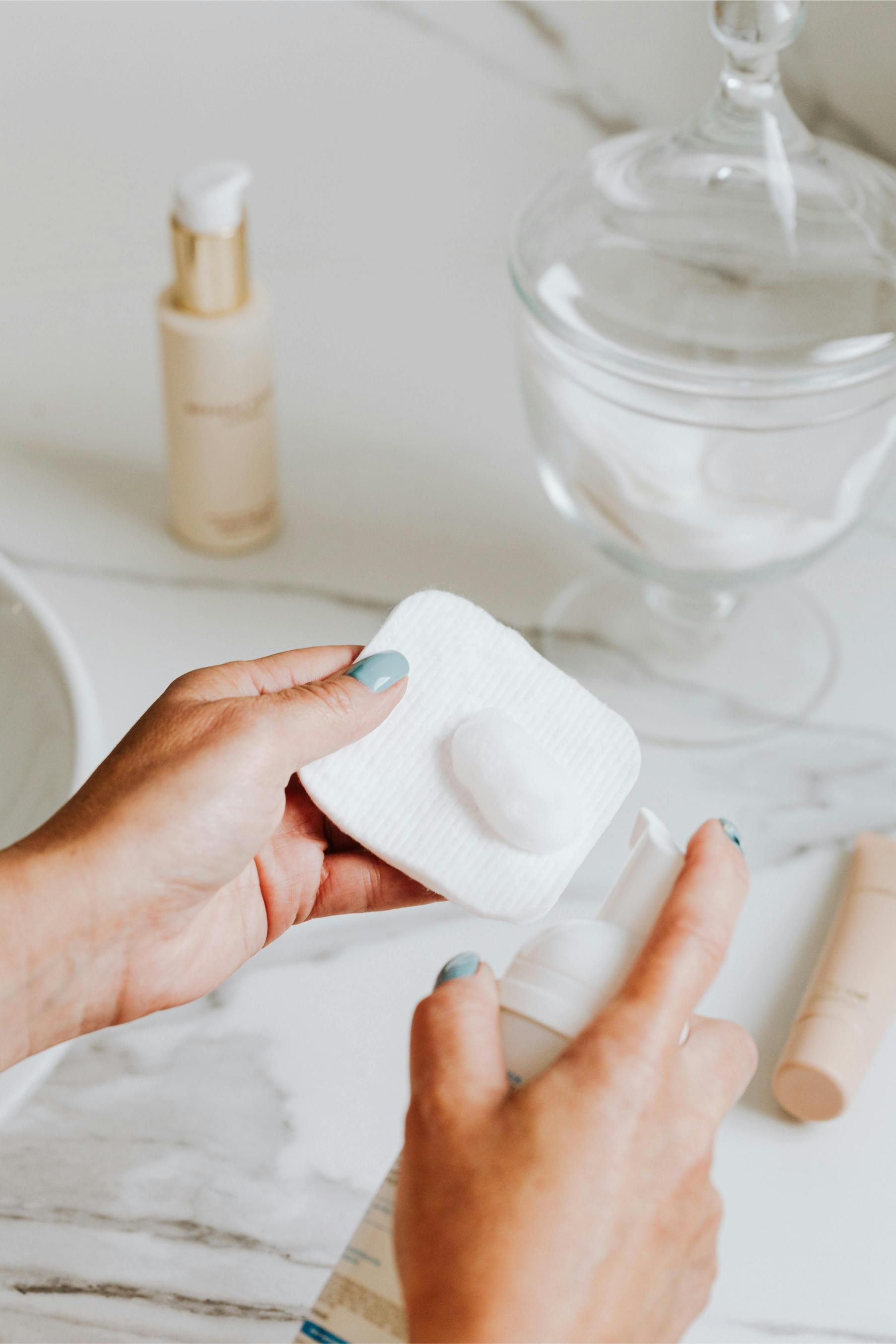 Hands applying a foaming cleanser to a cotton pad above a bathroom counter
