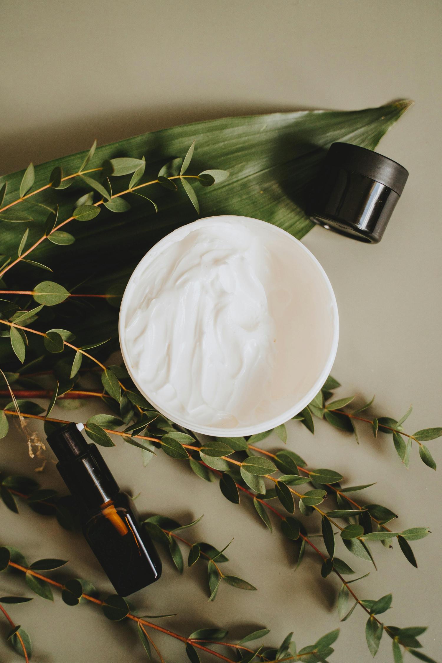 Tub of cream with amber skin care bottles next to it surrounded by greenery on a beige background