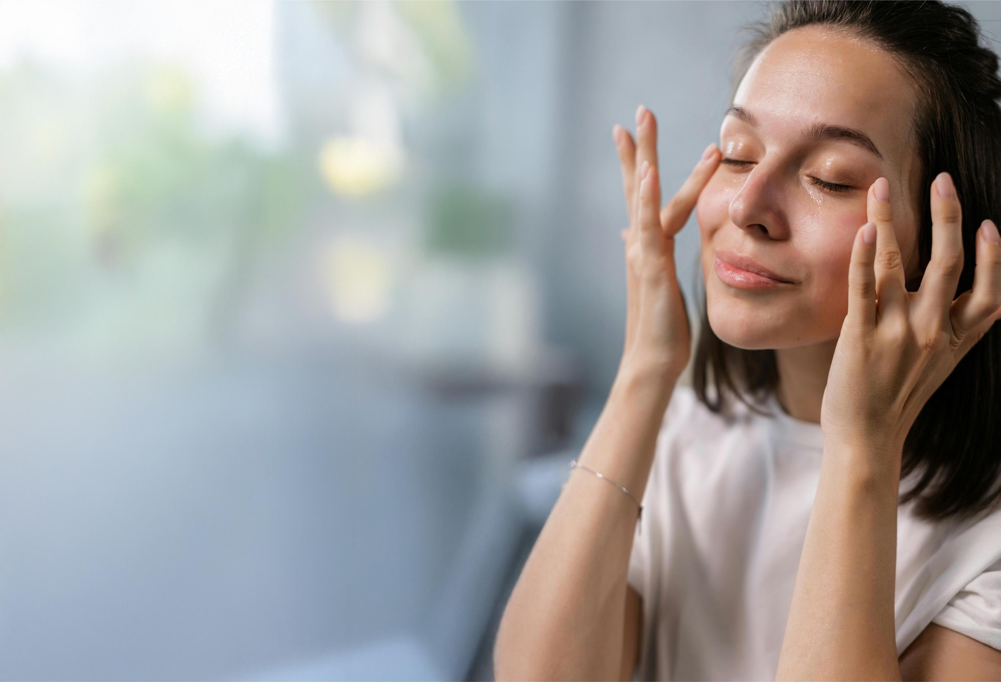 A woman gently massages her face while closing her eyes, appearing relaxed in a bright environment