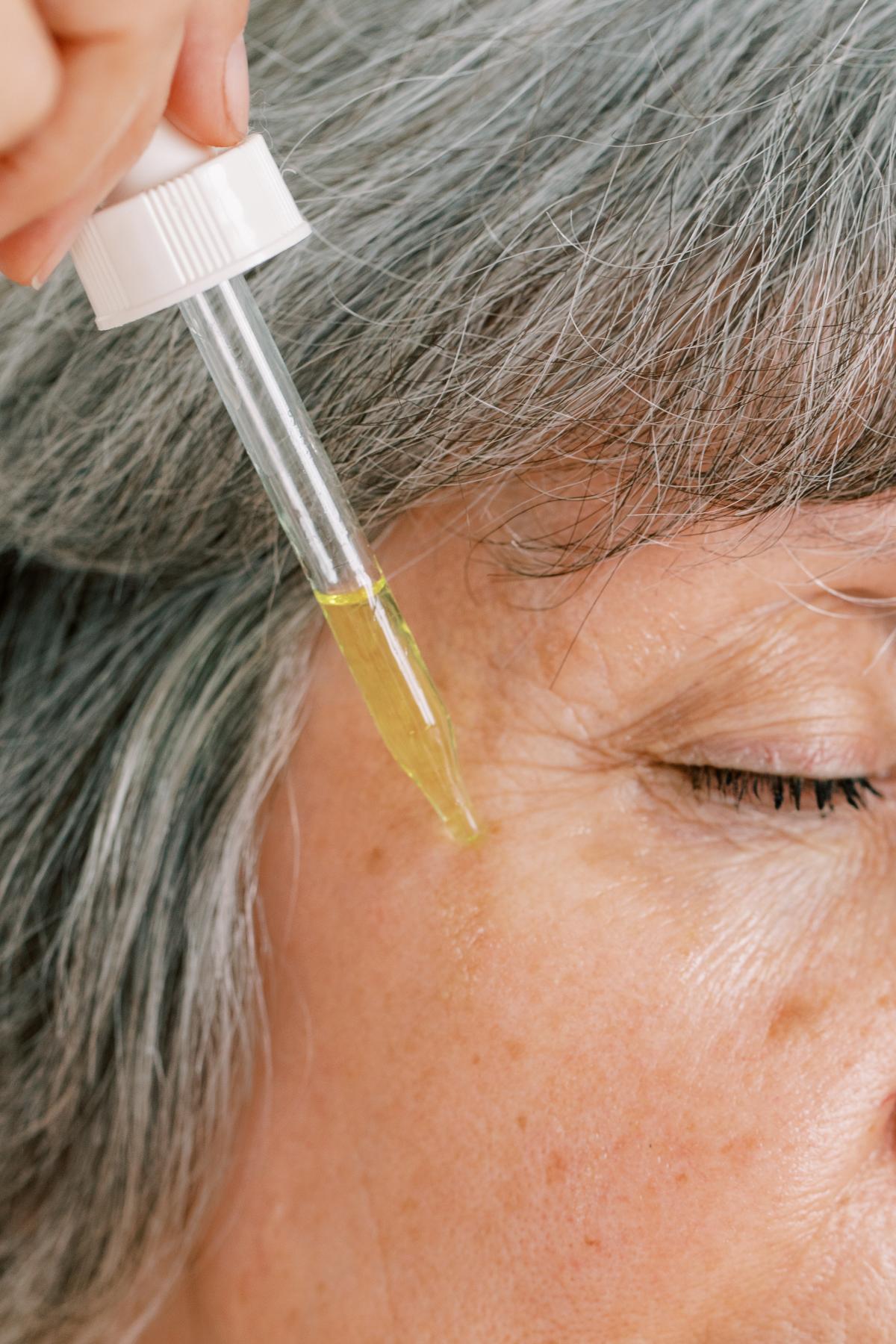 Close up of a woman with grey hair applying a yellow serum with a dropper to her face